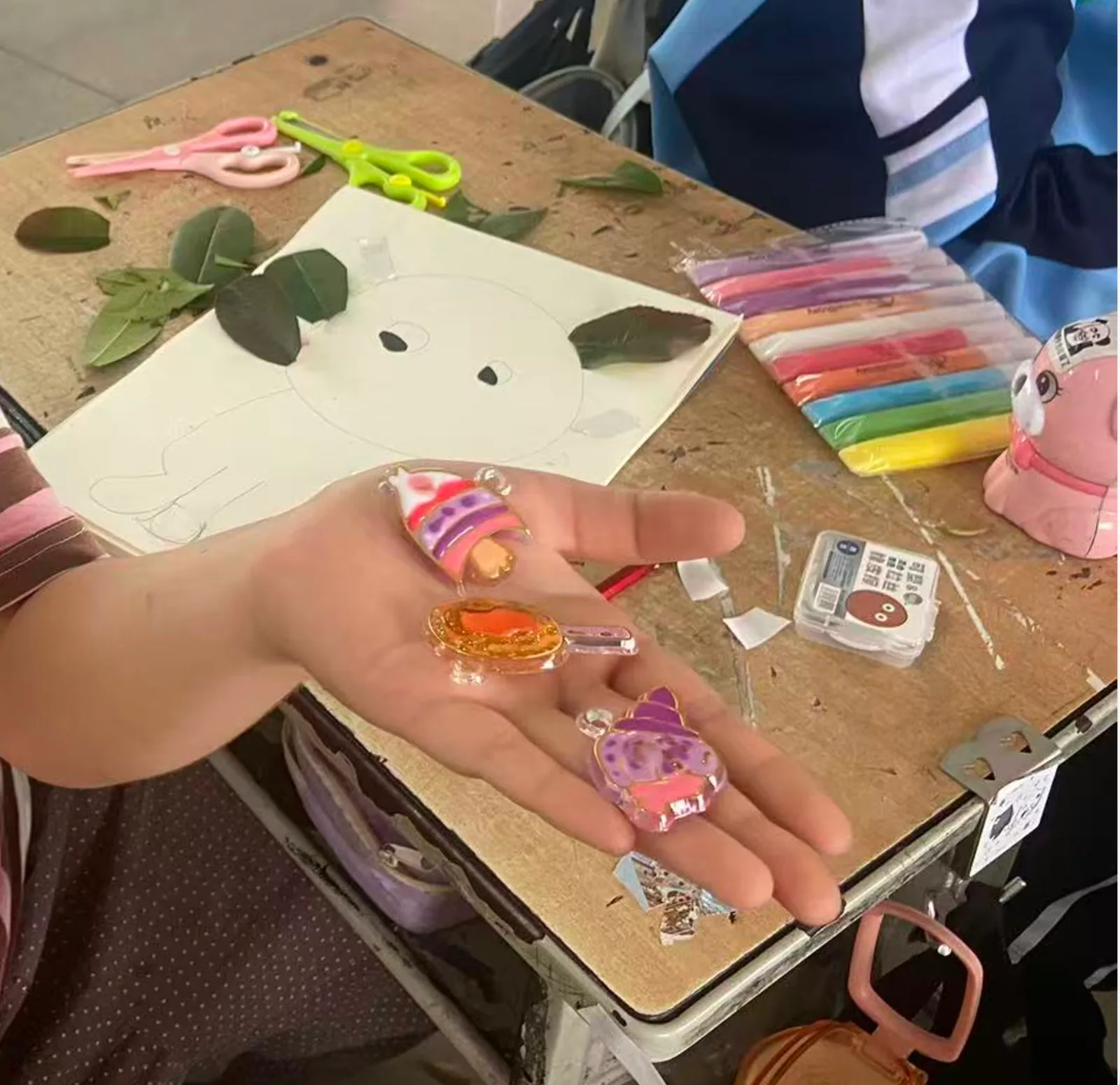 手心展示三个完成的彩色小胶画，桌上放着画笔和创作中的图案